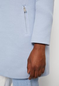 Svetle modrý kabát s herringbone texturou, s bočním zipem a volným střihem. Rukáv má manžetový design.