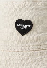 Chapeau seau beige en tissu texturé, présentant un patch en forme de cœur noir avec une broderie blanche "Carhartt WIP" sur le devant.
