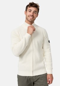 Strikket cremet cardigan med høj krave, lynlås foran og ribbede manchetter. Har struktureret stof og et lille sort logo på venstre ærme.