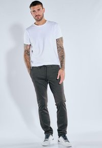 Mann mit tätowierten Armen, der ein weißes T-Shirt, dunkelgraue Jogginghosen und weiße Sneaker trägt, steht vor einem schlichten hellen Hintergrund.