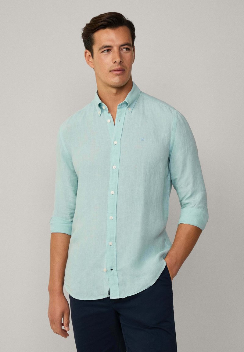 Camisa de lino azul claro con botones y mangas enrolladas, que presenta un cuello puntiagudo y un pequeño logo en el lado izquierdo del pecho, combinada con pantalones oscuros.