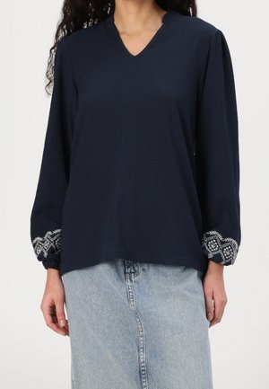 Kobieta ubrana w granatową bluzkę z długim rękawem, z białymi haftowanymi mankietami, oraz jasnoniebieską spódnicę jeansową.