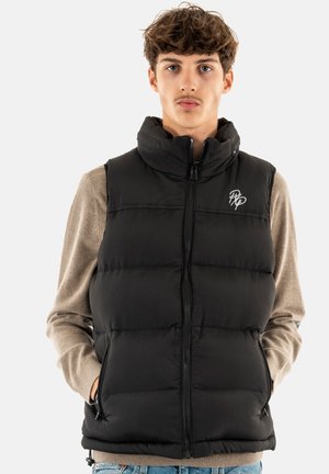 Gilet rembourré noir avec texture à côtes verticales, col montant, fermeture éclair avant, poches latérales et logo blanc sur la poitrine, porté sur un pull beige.