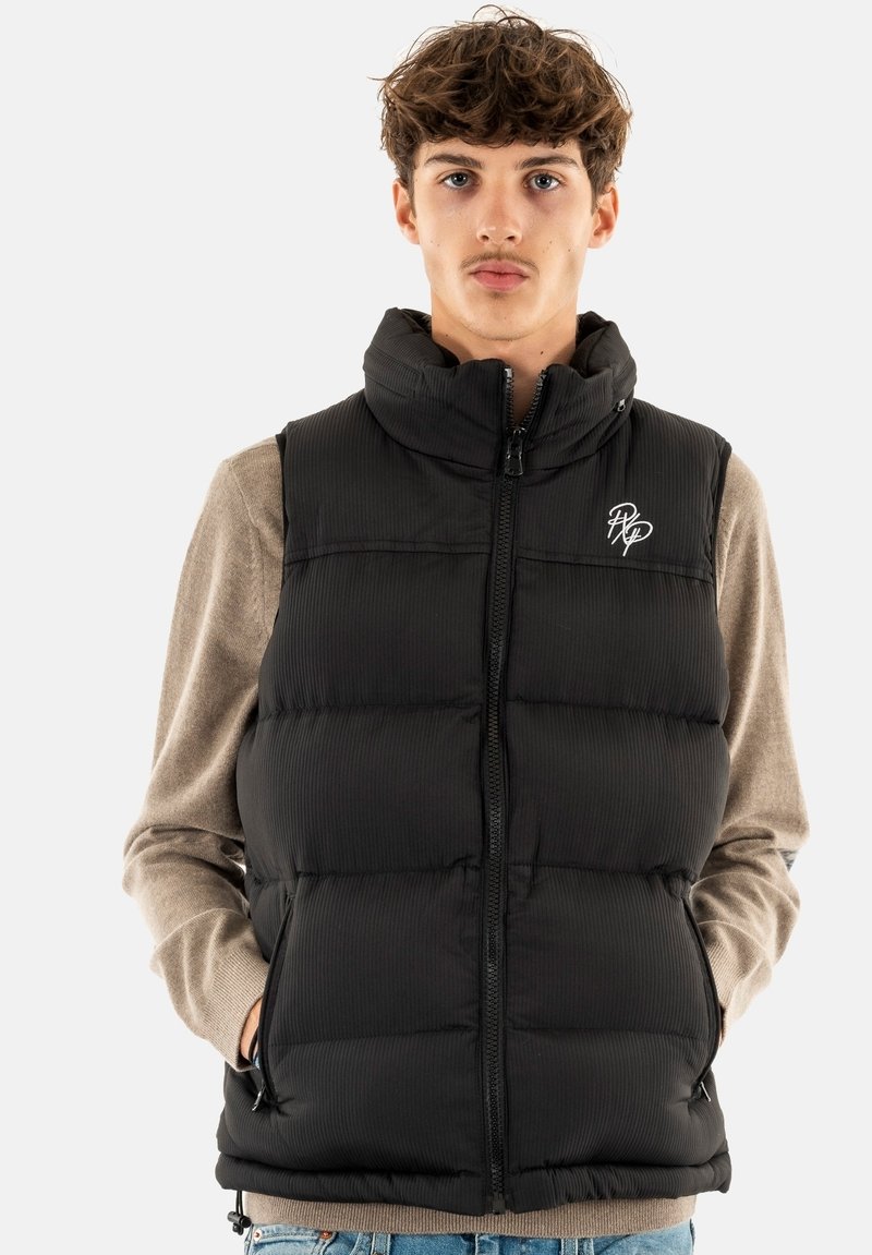 Chaleco acolchado negro con textura de canalé vertical, cuello alto, cremallera frontal, bolsillos laterales y logo blanco en el pecho, usado sobre un suéter beige.