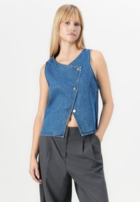 Denim mouwloos vest in een lichte blauwe was met een cropped ontwerp, voorzien van een unieke asymmetrische kraag en twee zilveren drukknoopjes.