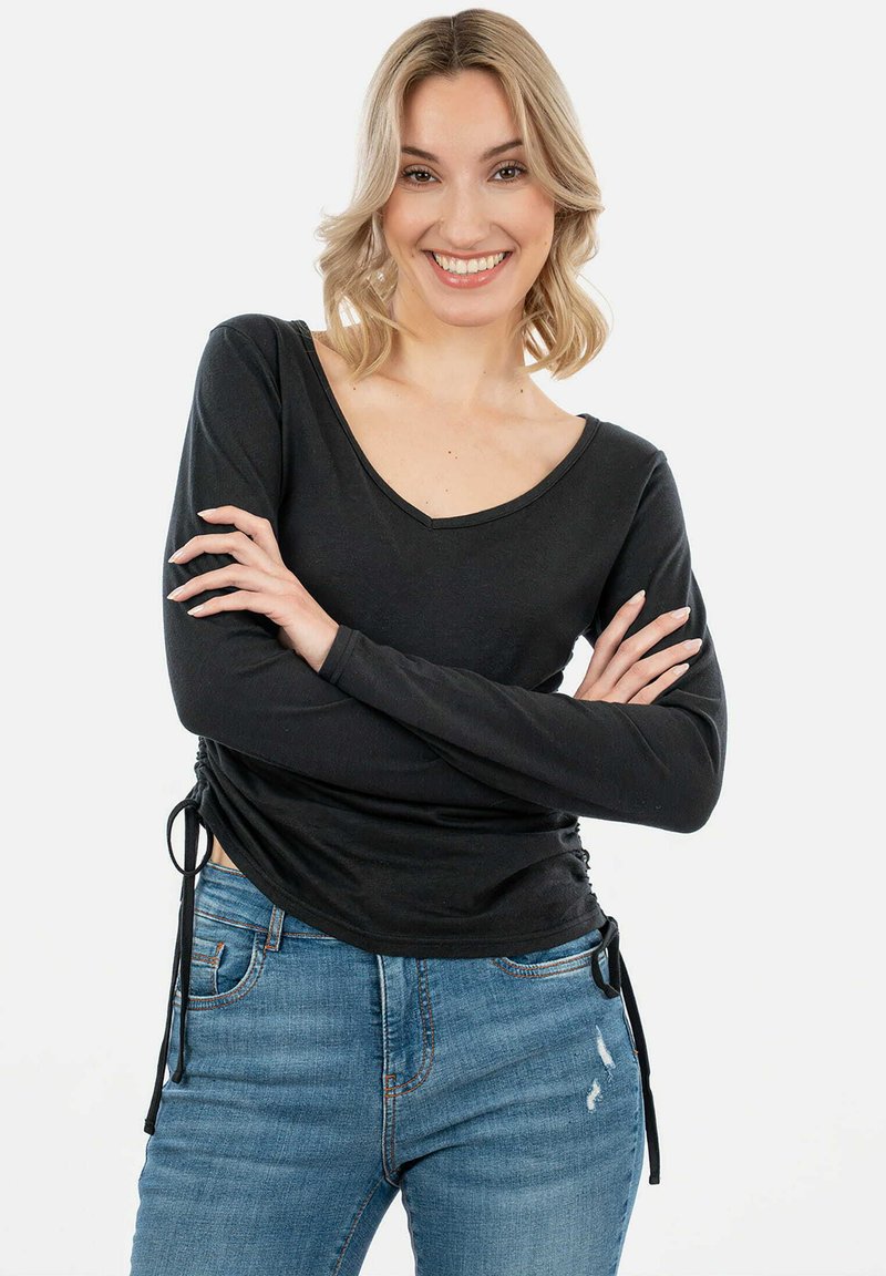 Must musta pikka varrukaga topp, millel on V-kaelus ja külgmised volditud detailid, koos helesiniste teksadega. Kangas näib olevat pehme ja elastne.