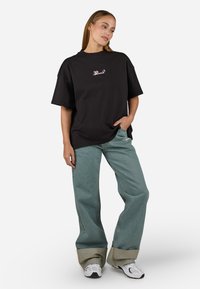 Black oversized t-shirt with front embroidery, paired with loose-fitting light blue jeans featuring contrasting green cuffs and white sneakers.