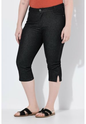 Frau trägt schwarze, kurz geschnittene Slim-Fit-Jeans mit Vordertaschen und kleinen seitlichen Schlitzen, kombiniert mit schwarzen Slip-On-Sandalen und einem orangen Oberteil.