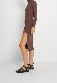 Vestito a maniche lunghe, a collo alto, rigato e marrone, con uno spacco alto, abbinato a sandali neri con cinturini e una chiusura regolabile.