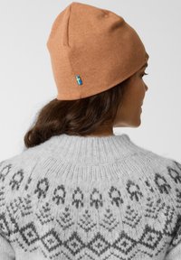 Beanie di maglia marrone con una vestibilità aderente, caratterizzato da un piccolo accento della bandiera svedese. La persona indossa un maglione grigio chiaro a fantasia.