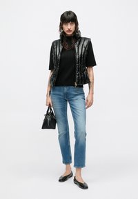 Čierna lesklá prešívaná vesta cez čierny pletený top, kombinovaná so svetlo modrými džínsami s rovnými nohavicami a čiernymi balerínami, držiaca malú čiernu kabelku.