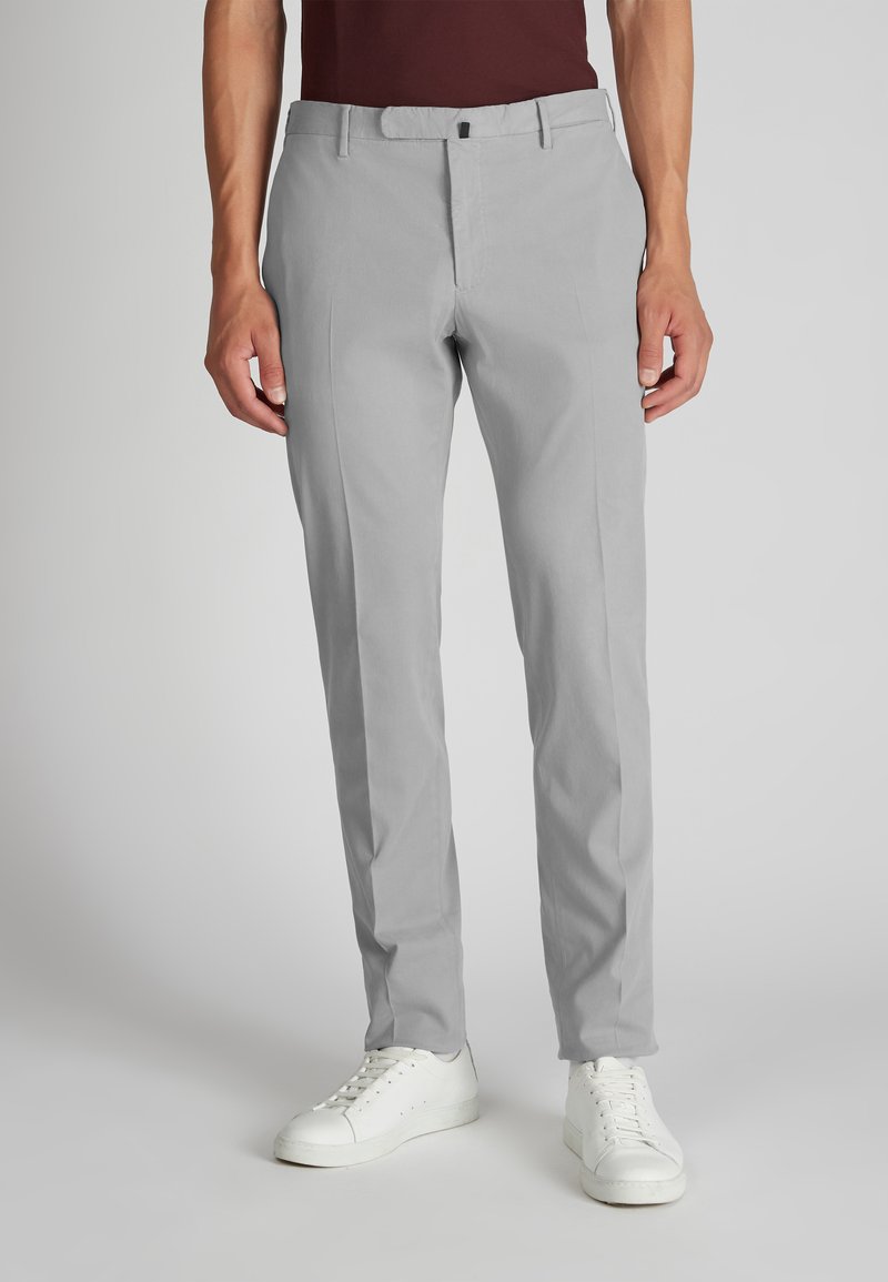 Mężczyzna w jasnoszarych, dopasowanych spodniach i białych trampkach, stojący na tle jednolitego jasnego tła.