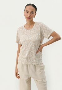 T-shirt beige à motifs floraux en tissu doux, avec un col rond et des manches courtes, porté avec un pantalon ample de couleur claire.