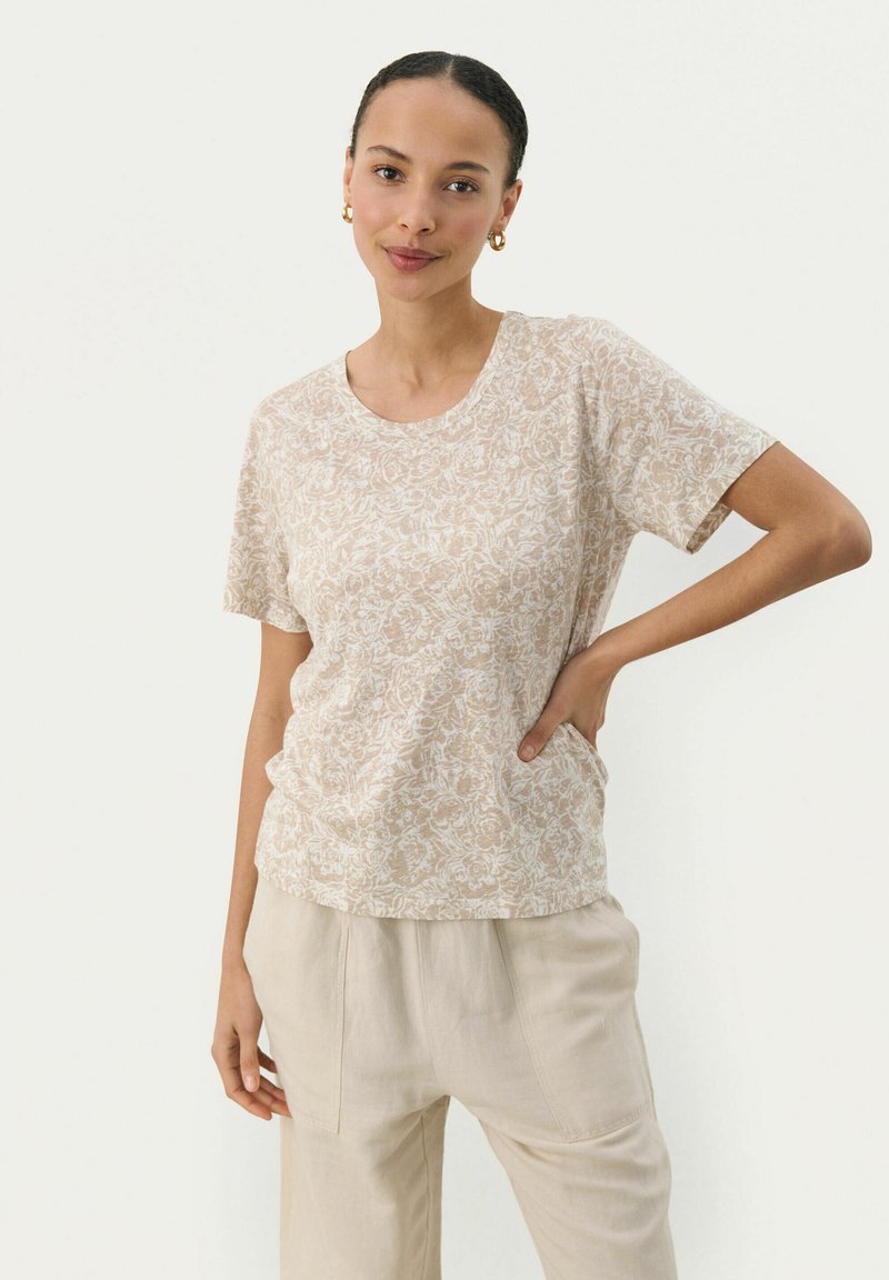 T-shirt beige à motifs floraux en tissu doux, avec un col rond et des manches courtes, porté avec un pantalon ample de couleur claire.