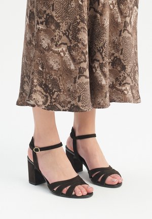 Sandali con tacco a blocco in suede nero, con cinturini alla caviglia regolabili e diverse cinghie incrociate sulla punta, su uno sfondo di un vestito con motivo pitone.