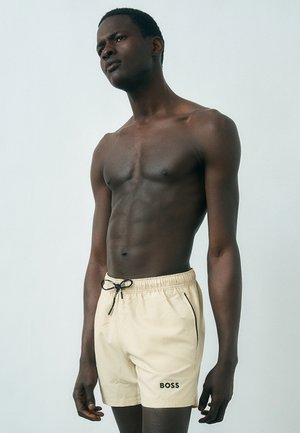 Muž bez trička s tmavou pokožkou, ktorý má na sebe béžové plavkové šortky BOSS s čiernou šnúrkou, stojaci pred jednofarebným svetlým pozadím.