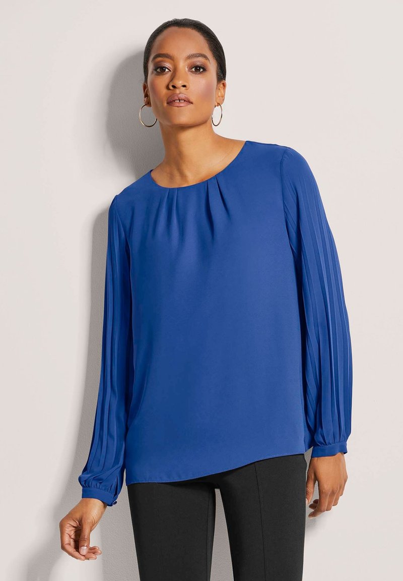 Blaue langärmelige Bluse mit Rundhalsausschnitt, die über plisseeartige Ärmel und eine lässige Passform verfügt. Glattes Material mit dezentem Schimmer.