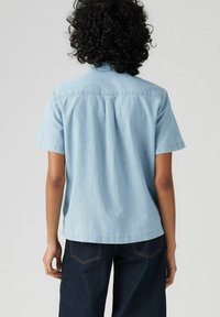 Light blue short-sleeve shirt made of soft fabric, featuring a collar and a back pleat. Paired with dark denim pants, showcasing a casual style.