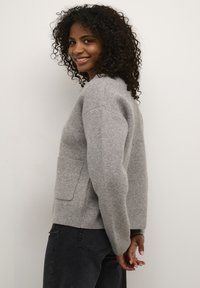 Maglione grigio in lana con scollatura arrotondata, polsini a coste e dettagli sulla tasca. Indossato sopra jeans scuri. Tessuto caldo e texture morbida.