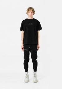 Camiseta negra de algodón con un estampado rectangular en el pecho, combinada con joggers negros que tienen detalles desgastados y bolsillos laterales con cremallera. Botas blancas.