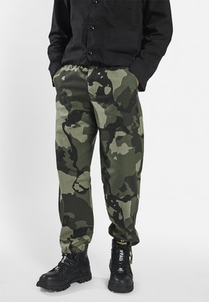 Camo pants in green and black hues, featuring a relaxed fit, elastic waist, and textured fabric. Paired with chunky black boots.