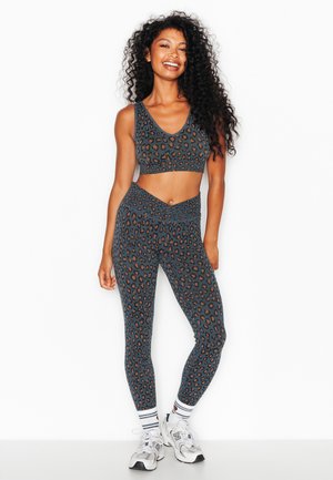 Donna sorridente che indossa un reggiseno sportivo e leggings coordinati con stampa leopardata scura, sneakers bianche e calzini bianchi a righe, in piedi su sfondo bianco.
