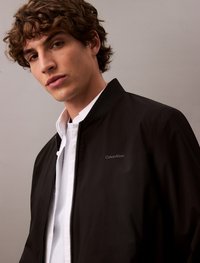 Black bomber jacket with zip closure and ribbed collar, featuring a small white "Calvin Klein" logo on the chest. Smooth fabric texture.