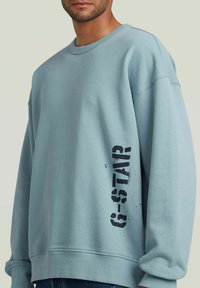 Sudadera azul claro con cuello redondo, puños acanalados y un ajuste ligeramente holgado. Presenta un estampado gráfico negro en el costado.