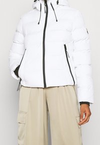 Hvid dunjakke med hætte, sort lynlåslukning og snoretræk. Har sidelommer og et let cropped design. Glat tekstur.