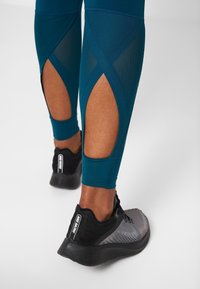 Turkoosi legginsit, joissa on verkko-leikkaukset sääressä, yhdistettyinä mustiin ja harmaisiin tennareihin, joissa on teksturoitu pinta ja pehmustettu pohja.