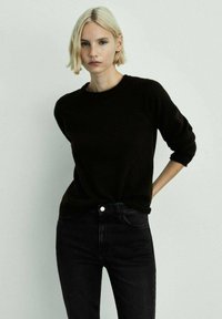 Černý svetr s kulatým výstřihem z měkkého materiálu, kombinovaný s černými džíny s vysokým pasem; modelka má krátké blond vlasy a stojí na světlém pozadí.