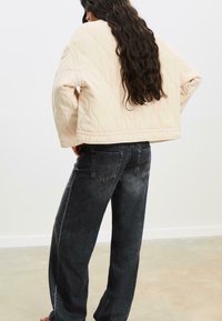 Quiltet beige jakke med en boxy pasform, der har lange ærmer og en cropped kant, parret med sorte denim jeans, som har baglommer.