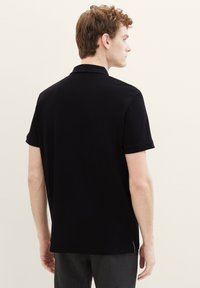 Zwarte polo met korte mouwen, standaardkraag en een zachte textuur. Bevat subtiele zijslits en heeft geen zichtbare logo's of patronen.