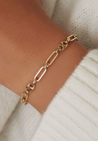 Bracelet en chaîne dorée avec des maillons allongés et circulaires au fini lisse, porté sur un poignet contre un fond en tissu texturé blanc.