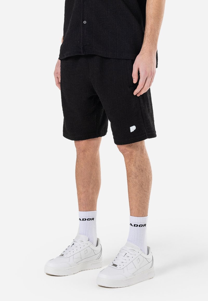 Personne portant un short noir texturé, des chaussettes blanches avec un texte noir et des baskets blanches, debout sur un fond blanc.