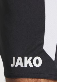 Juodi sportiniai šortai, pagaminti iš lygaus audinio, su balta šonine juosta ir žodžiu "JAKO" baltu tekstu ant apatinės kojos.