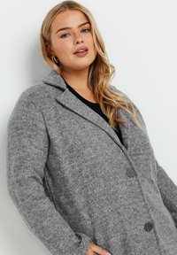 Grå kappa i ullblandning med en strukturerad design, stor krage, två knappar och sidofickor. Det texturerade tyget ger djup åt utseendet.