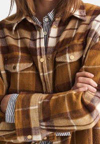 Camisa de franela a cuadros marrón y amarilla con cierre de botones y dos bolsillos en el pecho. Presenta herrajes en tono dorado y una camiseta de rayas debajo.