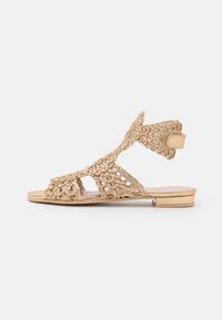 Guldöppen sandal med intrikat vävt mönster, platt klack, justerbar ankelrem och mjuk textur på en ljus bakgrund.