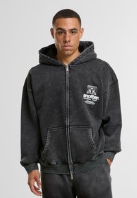 Dunkelgraues Zip-Hoodie mit einem verblassten Waschung, ausgestattet mit einem grafischen Druck auf der Vorderseite, einer Kängurutasche und gerippten Bündchen mit strukturierter Oberfläche.