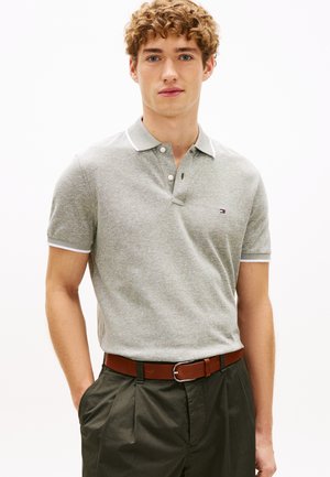 Młody mężczyzna o kręconych włosach, ubrany w jasnoszarą koszulkę polo, brązowy pasek i ciemnozielone spodnie, stojący z rękami w kieszeniach na białym tle.