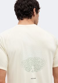 Άνδρας που φοράει ανοιχτό μπεζ μπλουζάκι Nike Running με κυκλικό μοτίβο από τελείες στο πάνω μέρος της πλάτης, φαίνεται από πίσω.