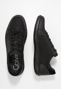 Schwarze Low-Top-Sneaker mit Schnürung, strukturierten Einsätzen und dicken Sohle, ein Schuh von oben und der andere von der Seite betrachtet.