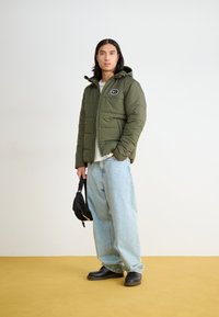 Žaliaveidis užpildytas džemperis su užtrauktuku ir kapišonu, su juoda logotipo etikete. Derinamas su laisvais šviesiai mėlynais džinsais ir juodais batais.