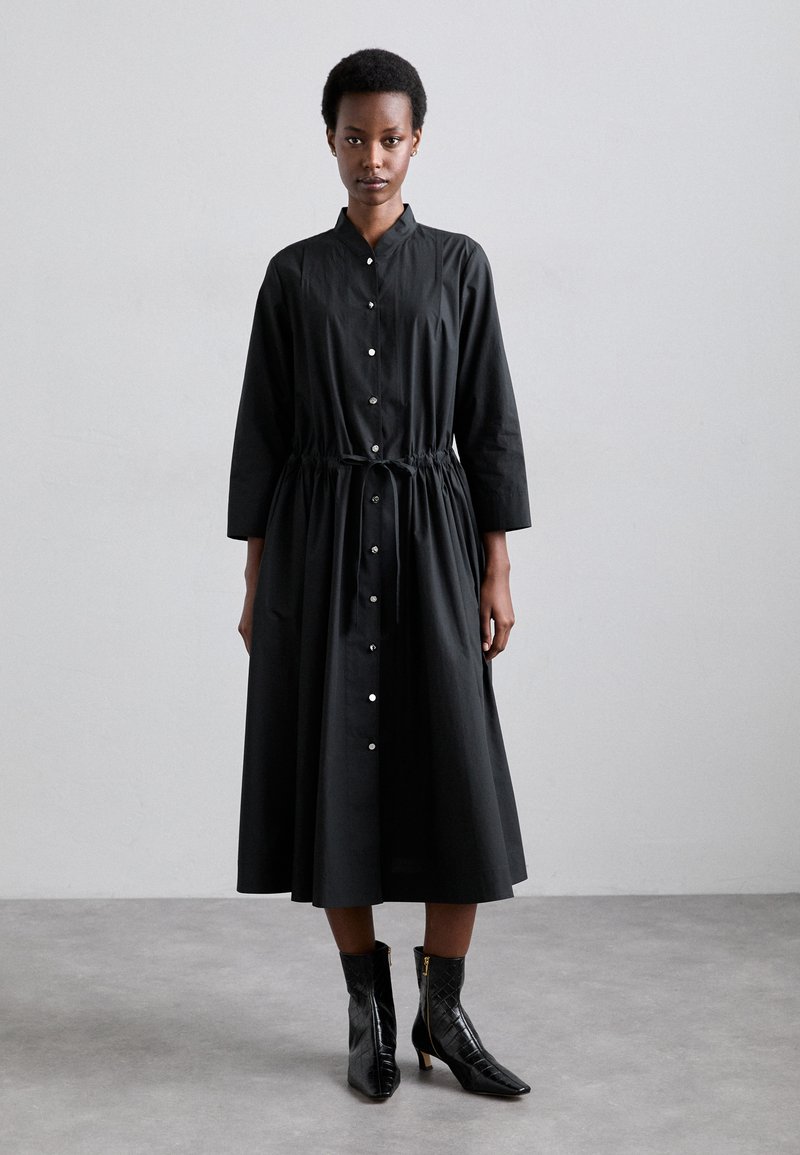 Vestido negro de manga larga con cierre de botones en la parte delantera, cintura ajustada y tejido suave. Combinado con botines negros brillantes con un diseño texturizado.