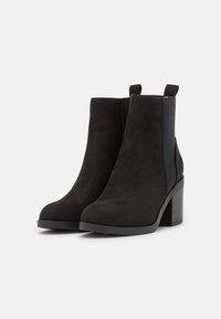 Bottines en suède noir avec un talon épais, dotées de panneaux latéraux élastiques et de tirants arrière pour un enfilage facile. Texture lisse avec un design simple.