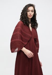 Burgunderfarget kjole med plissért stoff, dyp V-hals og brede, flytende ermer. Lett, strukturert materiale gir dimensjon til designet.