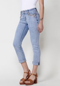 Svijetloplavi traperice s ukrasnim draguljima na džepovima, slim fit i podvrnutim manžetama. Uparene s tamnim sandalama na drvenom petu.