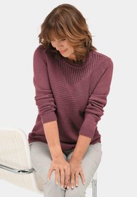 Strickpullover in Mauve mit strukturierten geometrischen Mustern und gerippten Bündchen. Das Model sitzt, die Hände auf den Knien, in hellgrauen Hosen.