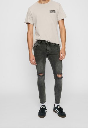 Mężczyzna w beżowej koszulce Urban Classics, podartych czarnych obcisłych dżinsach, białych skarpetkach i czarnych butach sportowych Adidas stoi na jednolitym tle.